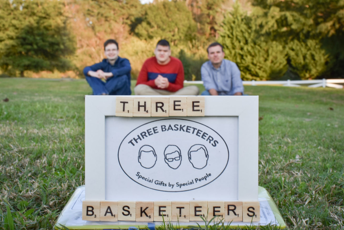 Close up of the Basketeers letter tile frame on the grass with three Basketeers sitting on the ground behind the frame