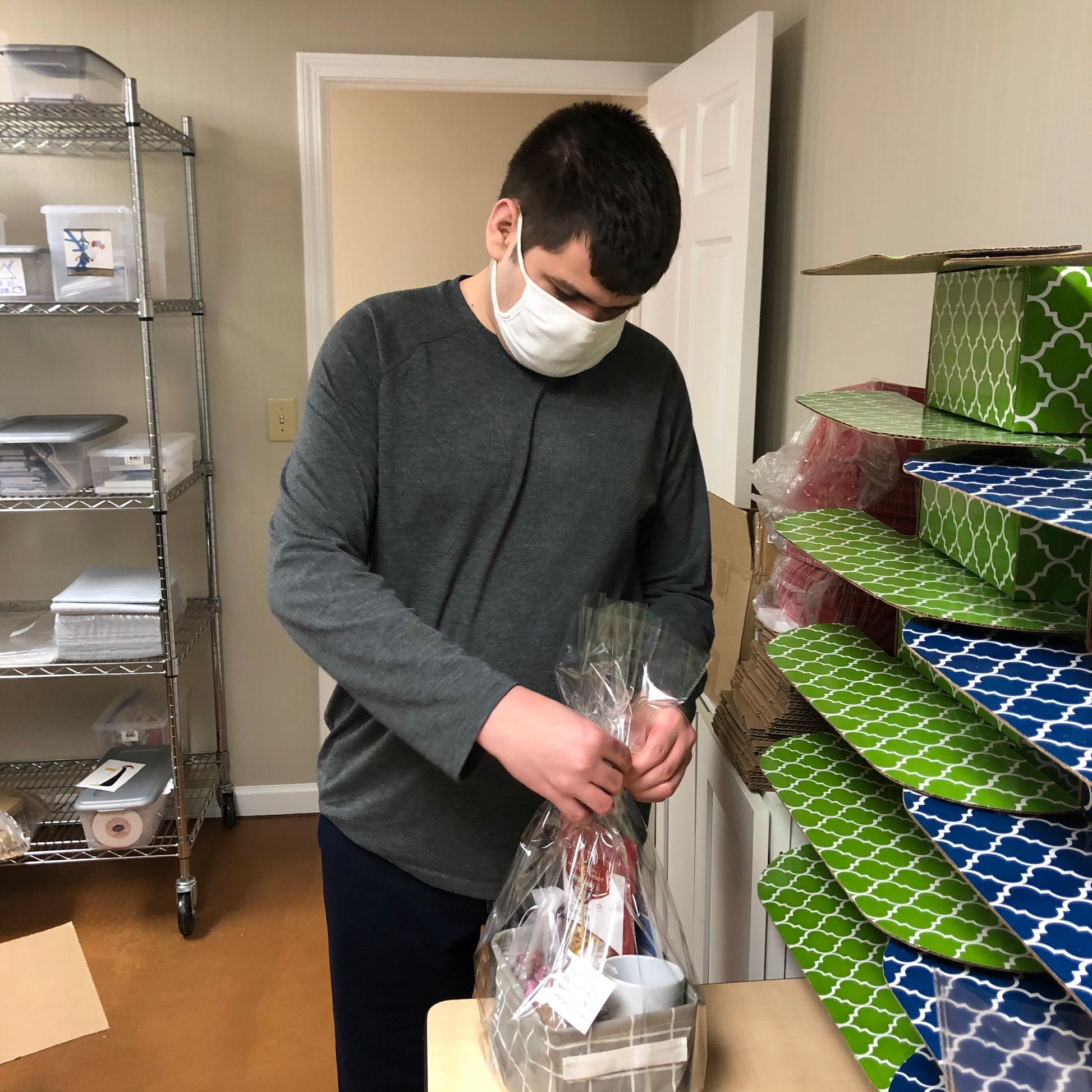 A disabled business owner with a gray shirt tying a ribbon on a "Just Because" gift basket while standing in an inventory room