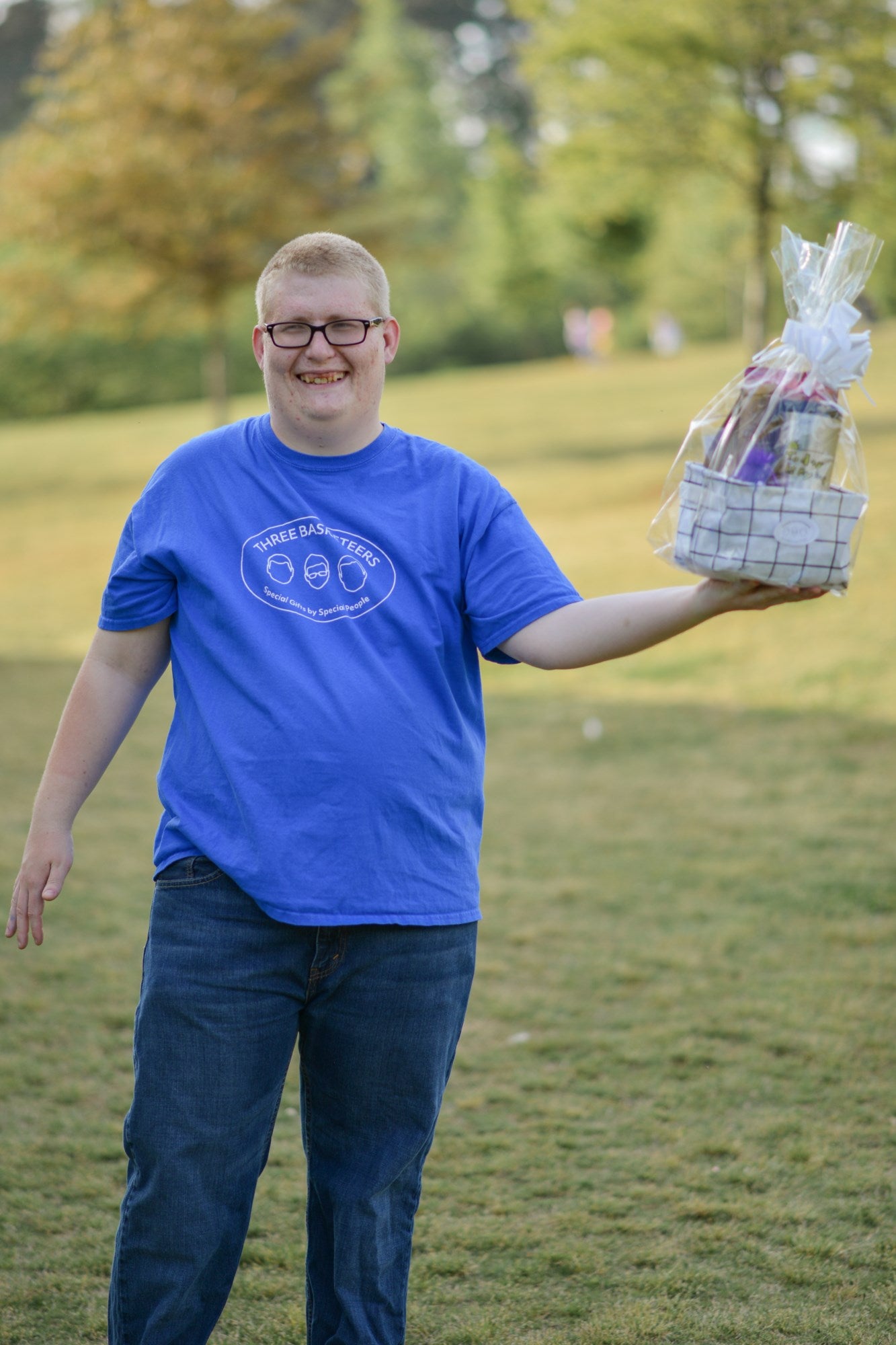 A disabled business owner with a blue shirt and glasses holding a completed "Just Because" gift basket in one hand while standing outside