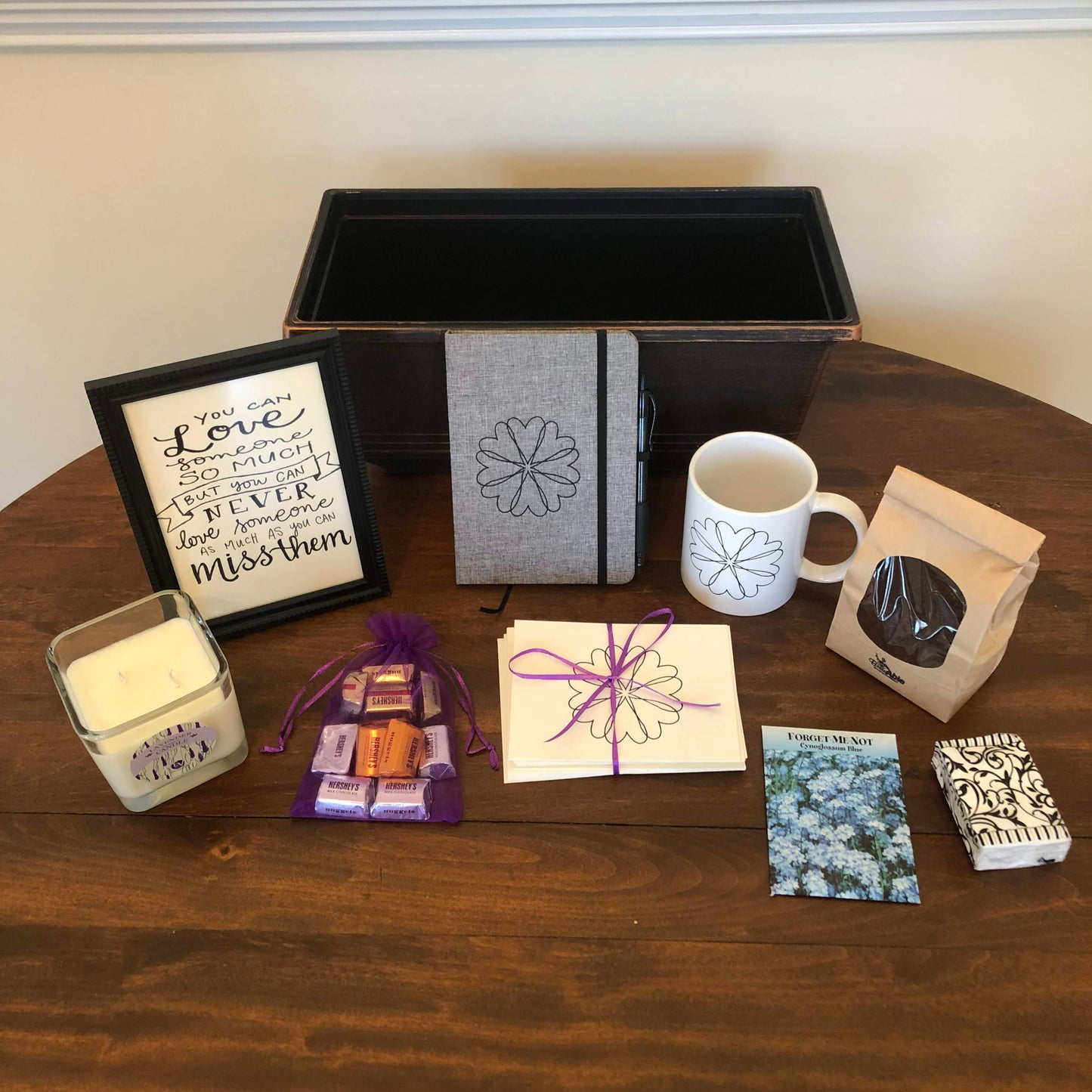 This condolence gift basket with upgraded wood planter includes a double wick lavender candle, handmade heart wreath notecards, gourmet coffee,  chocolate, ceramic mug, framed inspirational calligraphy, fabric covered journal, tissues and a forget me-not-seed packet