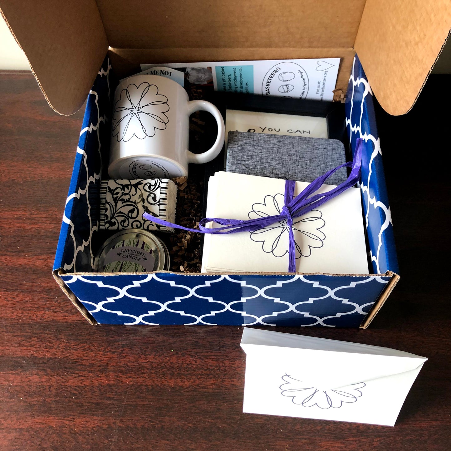 This condolence gift box includes a double wick lavender candle, handmade heart wreath notecards, gourmet coffee,  chocolate, ceramic mug, framed inspirational calligraphy, fabric covered journal, tissues and a forget me-not-seed packet in a patterned box ideal for shipping nationally