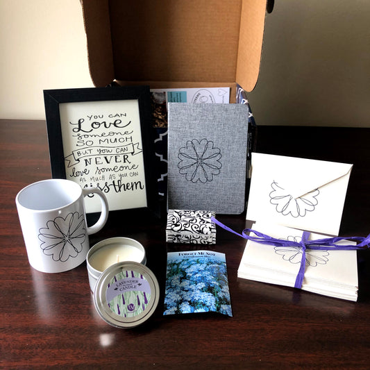 This condolence gift box includes a double wick lavender candle, handmade heart wreath notecards, gourmet coffee,  chocolate, ceramic mug, framed inspirational calligraphy, fabric covered journal, tissues and a forget me-not-seed packet in a patterned box ideal for shipping nationally